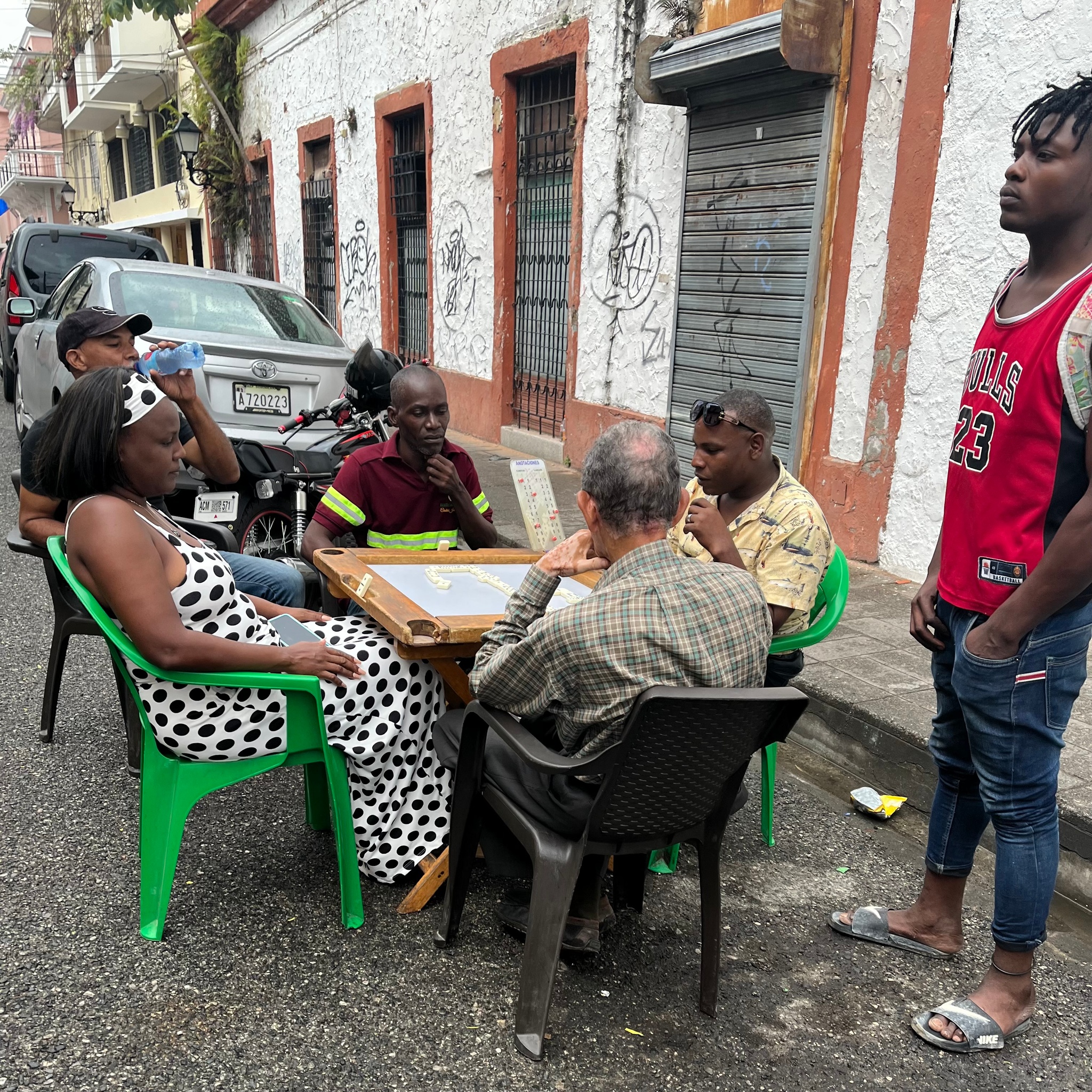 Six people gather around a board game that they are playing on a side street in a Dominican Republic neighborhood. Five of the players are seated around the table with their attention on the game. A sixth person stands to the side, looking over and away from the table. He wears a red Chicago Bulls jersey, blue pants, sandals, and has dark brown skin. 