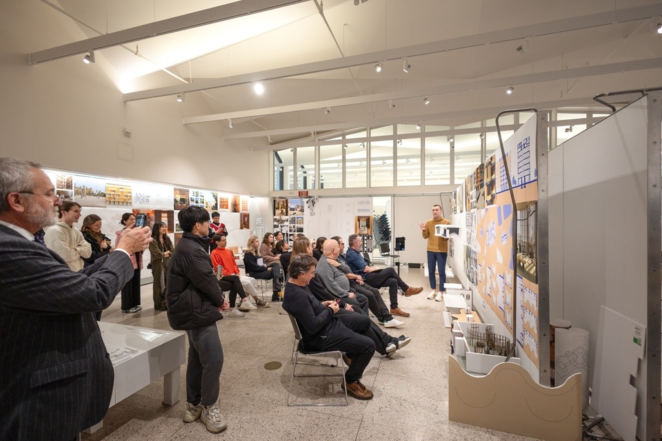 Wide shot of a gallery space set up with architecture student work. A student presents to a room full of faculty, critics, and classmates.