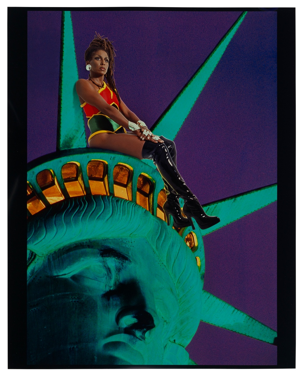 A photograph of a dark-skinned woman in a red, yellow, and green leotard and thigh-high black boots sitting on top of the head of the statue of liberty in front of a deep blue sky.