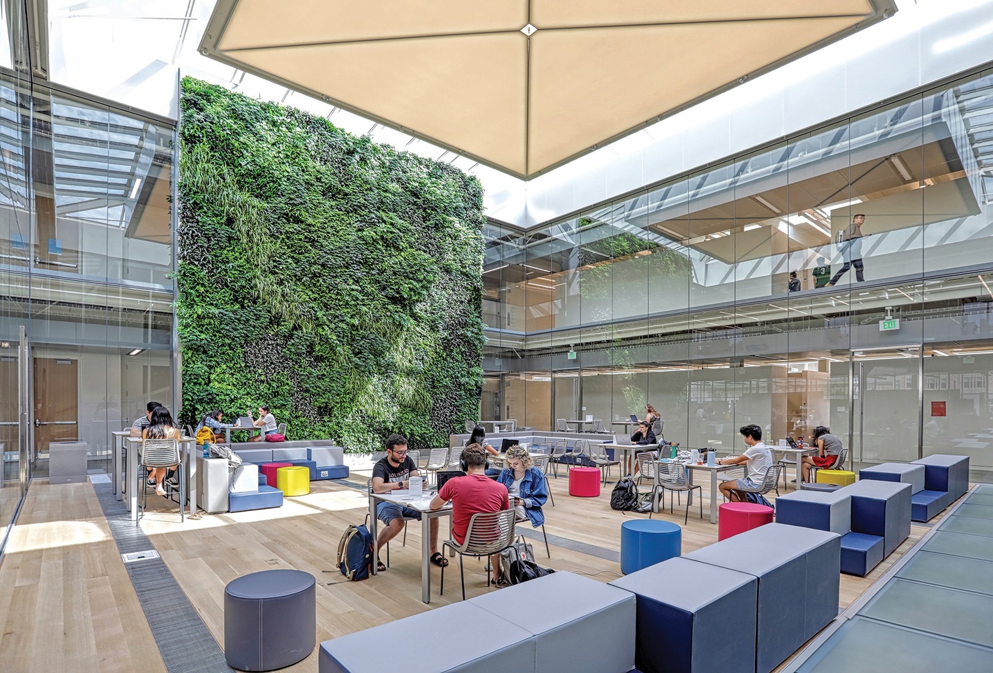 Light-filled, glass-walled court with people sitting at tables and around colorful soft seating; a two-story green wall filled with plants lines the back wall.