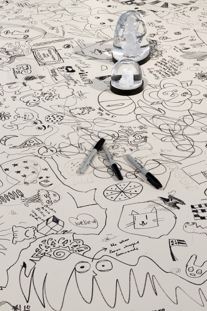 A different view of a white surface featuring black drawings and doodles, along with various blurbs of handwritten text, atop which sits two snow globes and three sharpies.