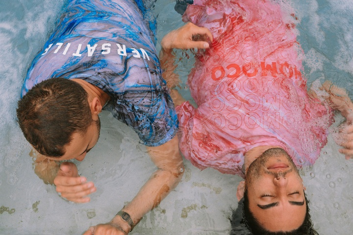 Two people lying in the sand with water running over them, one upside down face down and wearing a wet blue shirt, the other on their back face with a wet red/pink shirt with the word HOMOCO on the front.