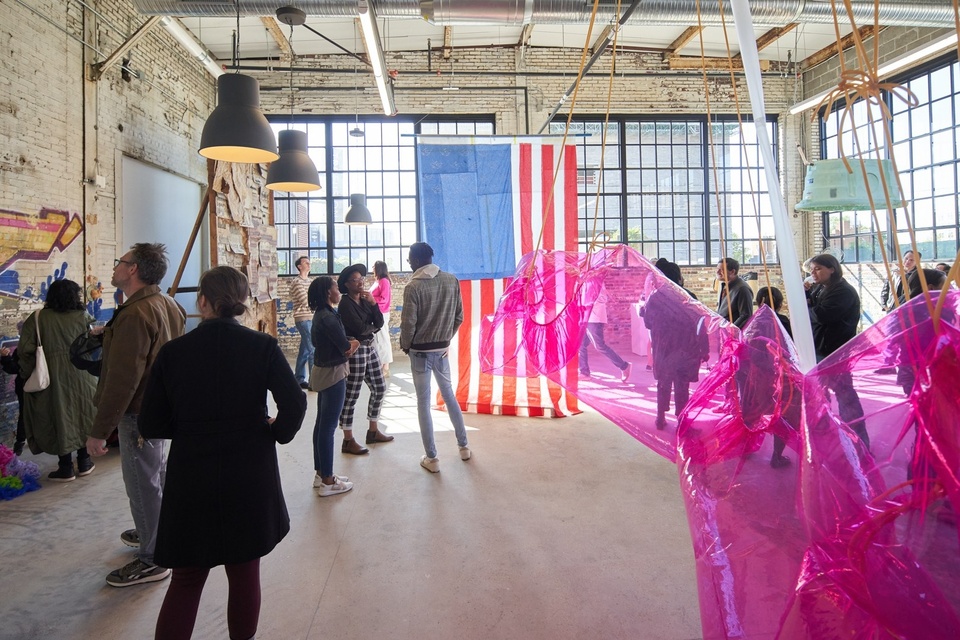 Raw warehouse space with floor to ceiling windows, concrete floors, and graffitied brick walls that is serving as a gallery space for an art exhibition. In the foreground is a pink plastic bubble-like sculpture, and in the background is hung a massive US flag with the blue section imprinted with a pattern that is not stars.