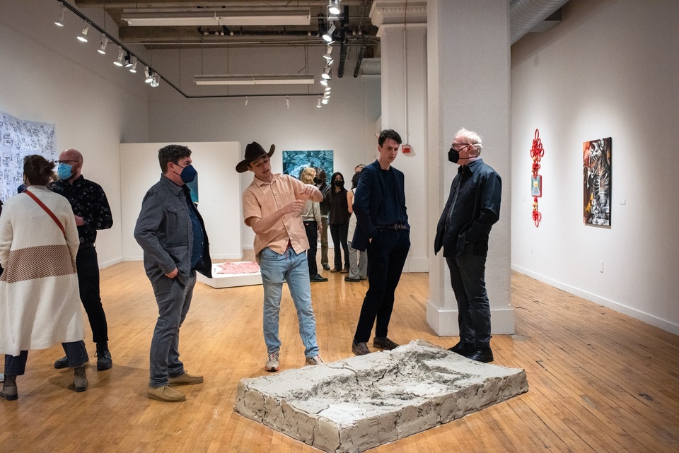 Several people stand in a gallery space around a rectangular sheet of concrete on the floor about the size of a tabletop with an impression of a small horse in it.