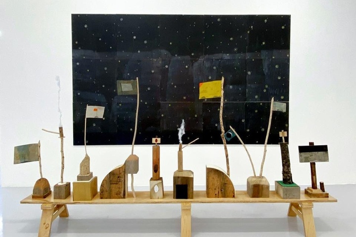Artwork installation by Tom Reed featuring several wooden sculptures, several with flags, on a wooden bench in front of a large rectangular black artwork with dots on the wall.