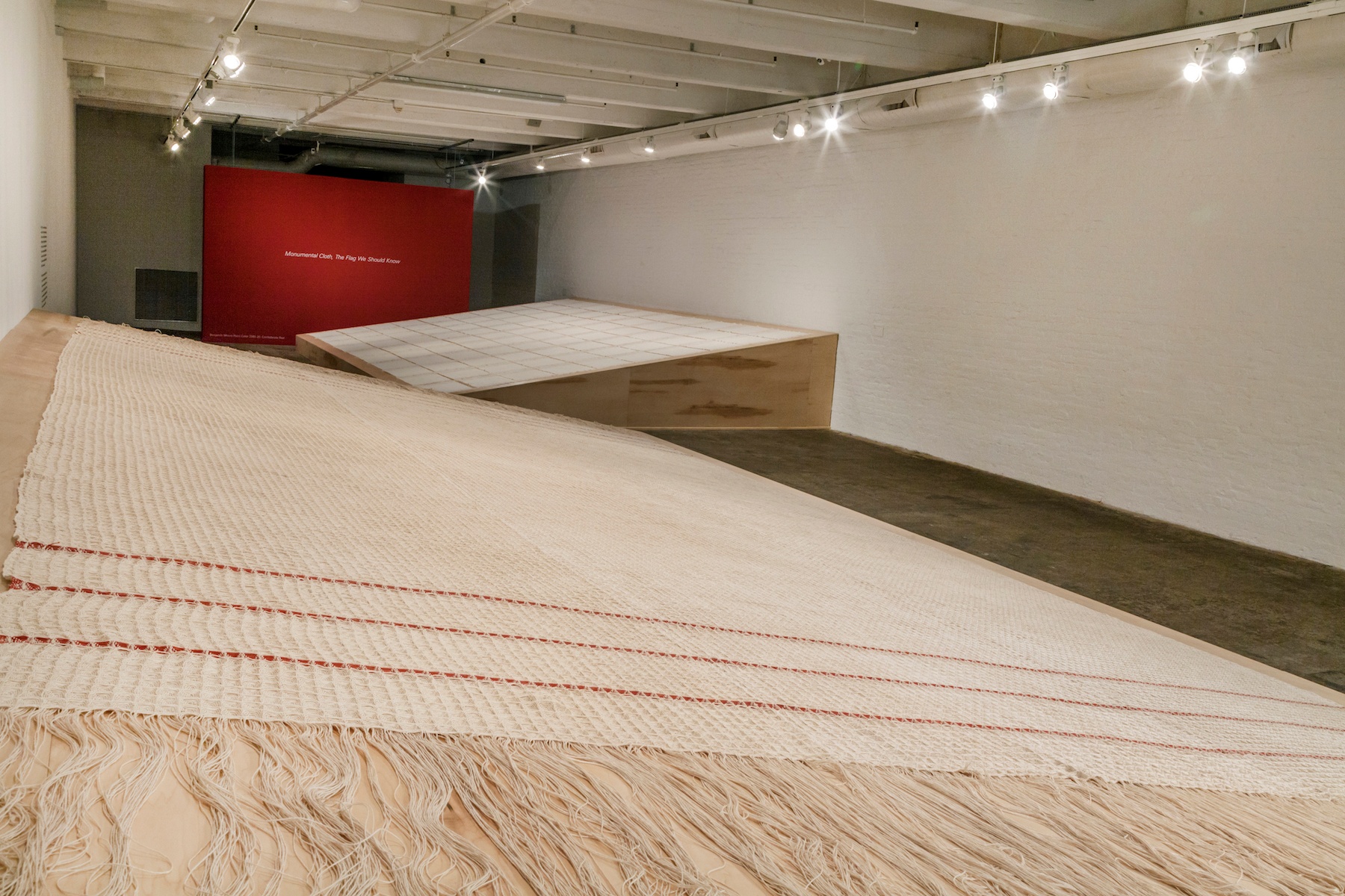 A woven cream textile with three red stripes on either end laying on a slanted wooden platform.