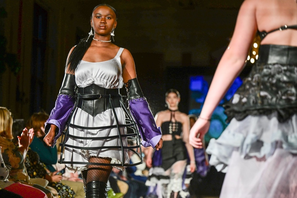 Models walk a fashion show runway in a collection of size-adjustable leather and lace dress combinations with purple accents.