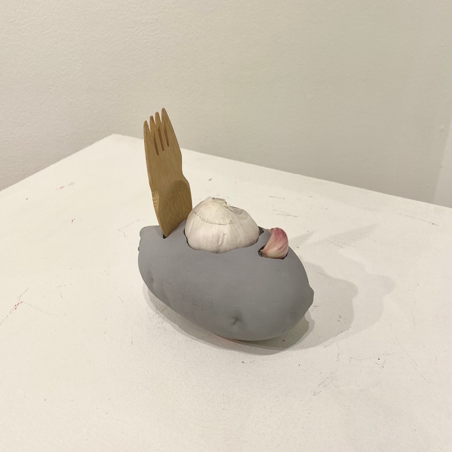 A sculpture comprised of a serving fork. an onion, and a clove of garlic all laid onto a round of clay. 