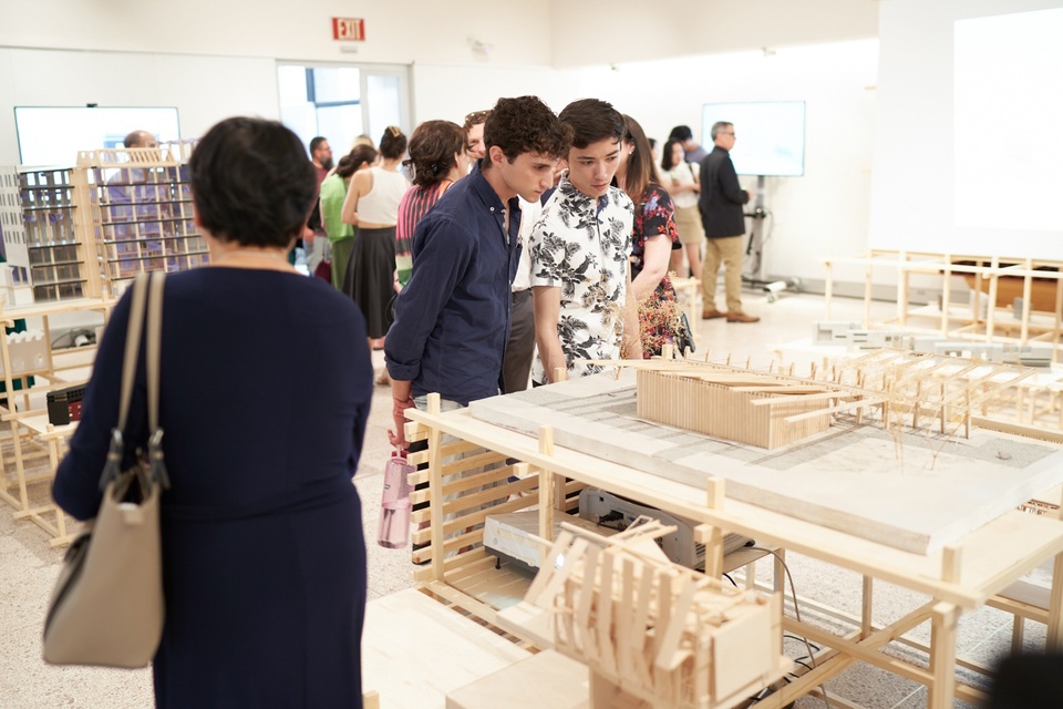 Two students lean over a wooden building model on a table in a gallery space. Other models are on display around the space and a crowd of student and parent attendees view the work.