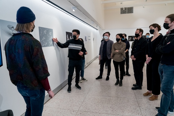 Person stands in a gallery space in front of a large crowd and points to a poster print of a digital architectural rendering pinned to the wall.