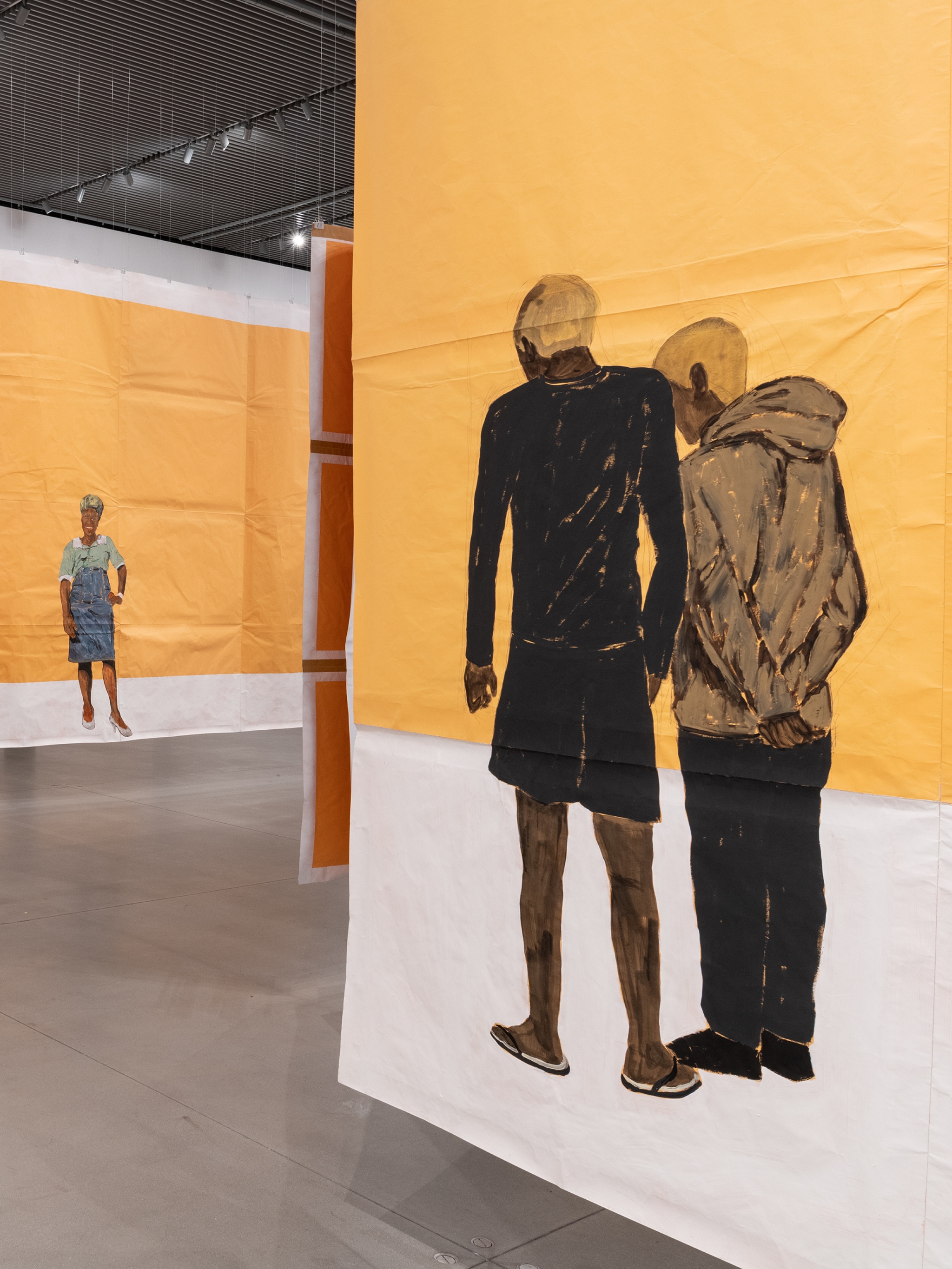An art gallery with paintings on paper that hang from the ceiling to form passages. A corner formed by paintings reveals a painting of a Black woman posing against a yellow brown background. Another painting in the foreground shows two young Black men standing together with backs to the viewer. They both have blond hair.
