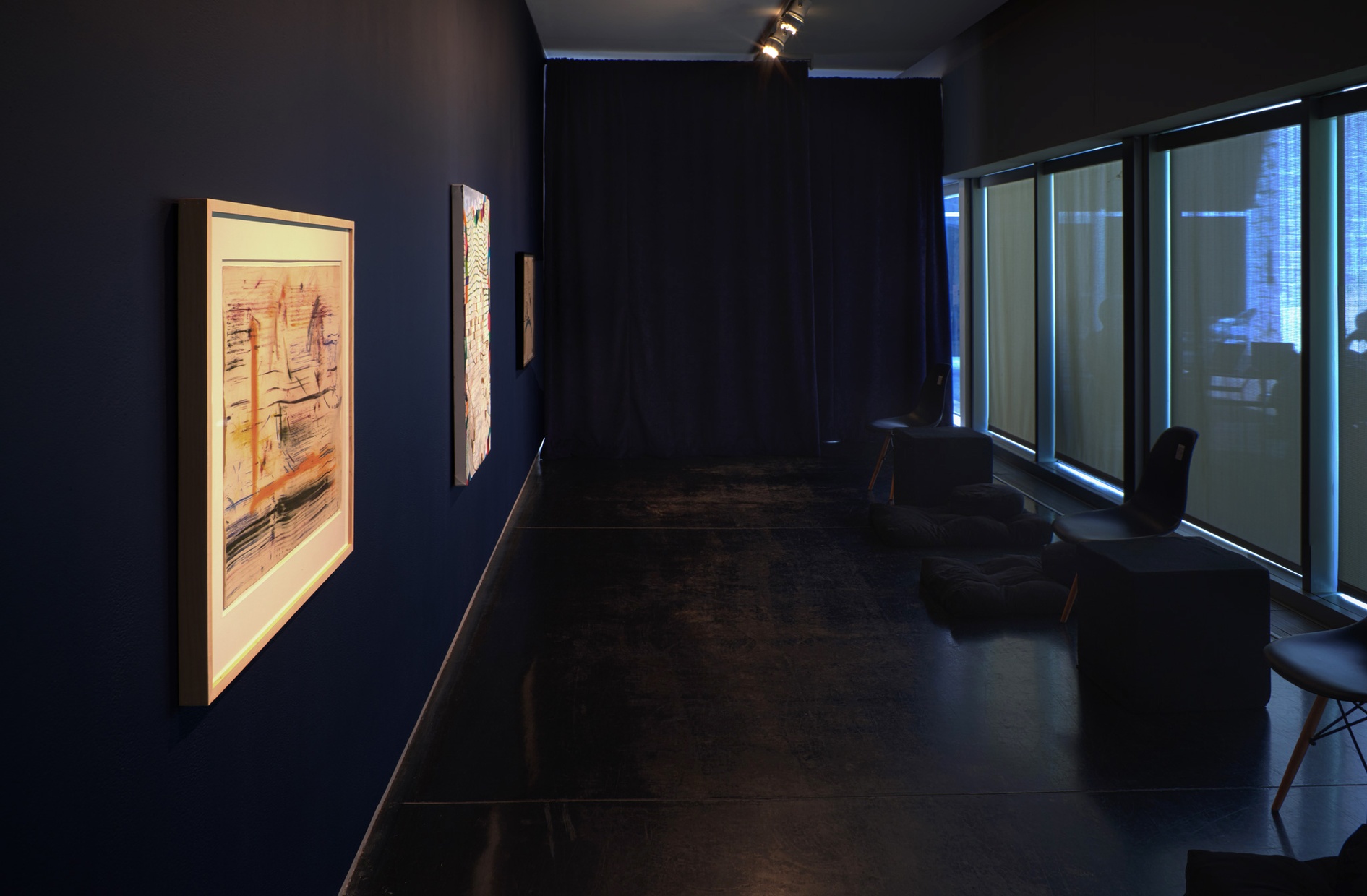 In a dark room with blinds covering the windows are multiple black chairs facing three abstract artworks against a blue wall.