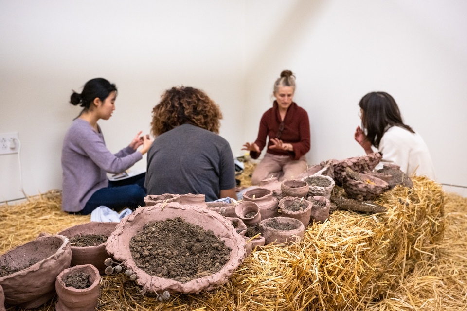 Close up of clay pots filled with a dirt-like substance sitting on a hay bale in an artist's studio. In the background four people sit on the floor.