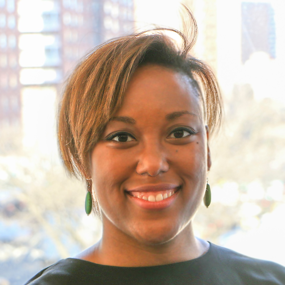 A Black woman with light brown, short hair styled at an angle across her forehead smiles. In the background is bright sunlight and city buildings. 