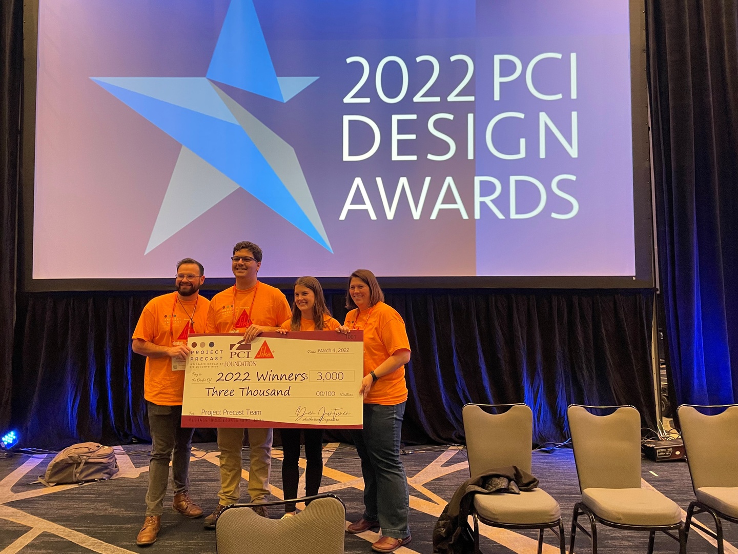 Photo of four team members in orange shirts holding an oversized check, in front of a digital screen for 2022 PCI Design Awards
