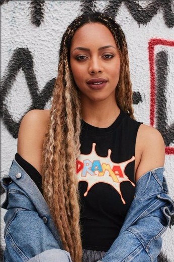 A Black woman with long hair stands against a gray wall with spraypainted writing on it. She wears a jean jacket pulled halfway down her arms.