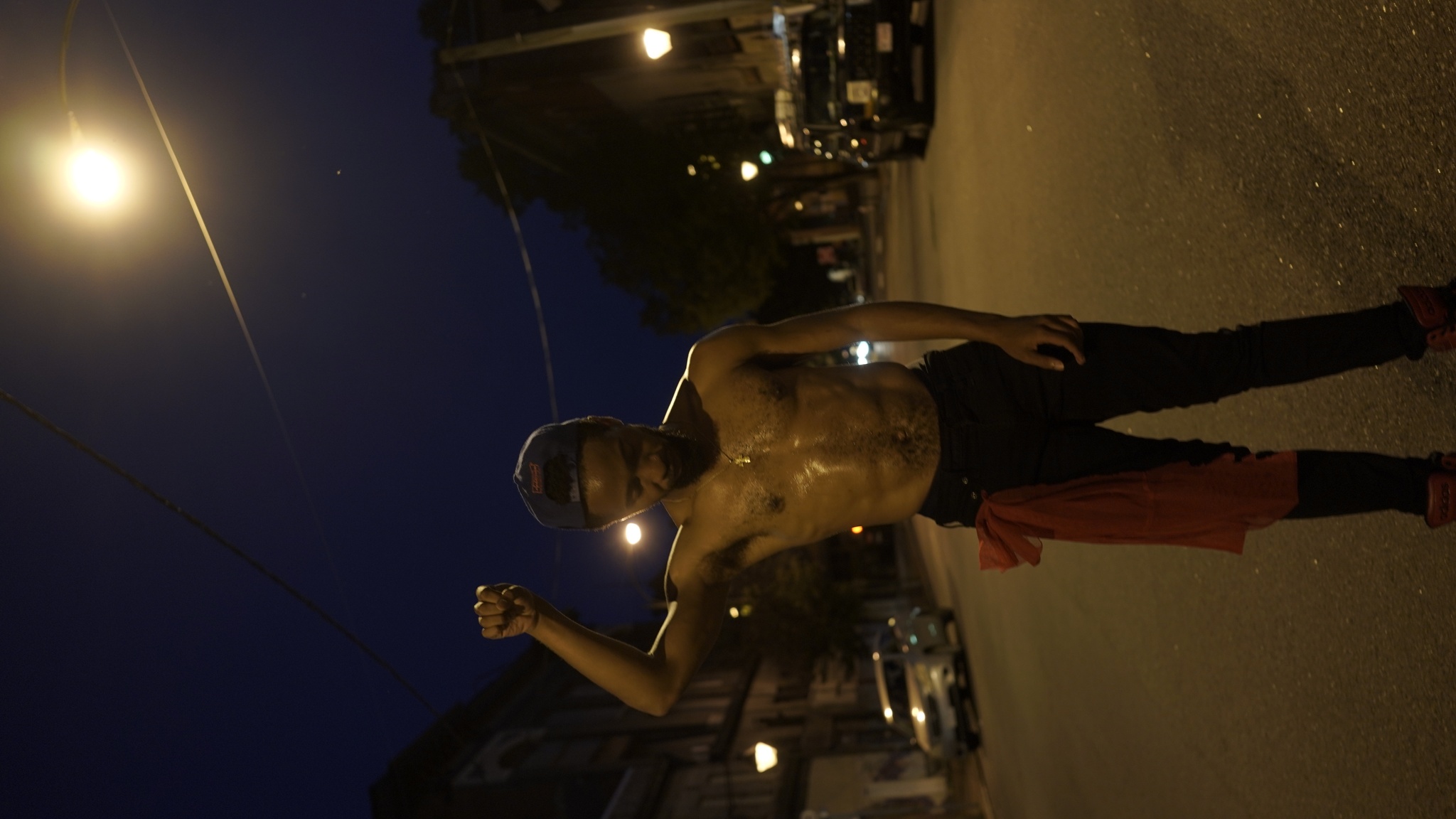 A Black man stands shirtless in a street at night, wearing a baseball cap backwards. He is sweaty as if he has just been exercising and holds his left fist up in the air. 