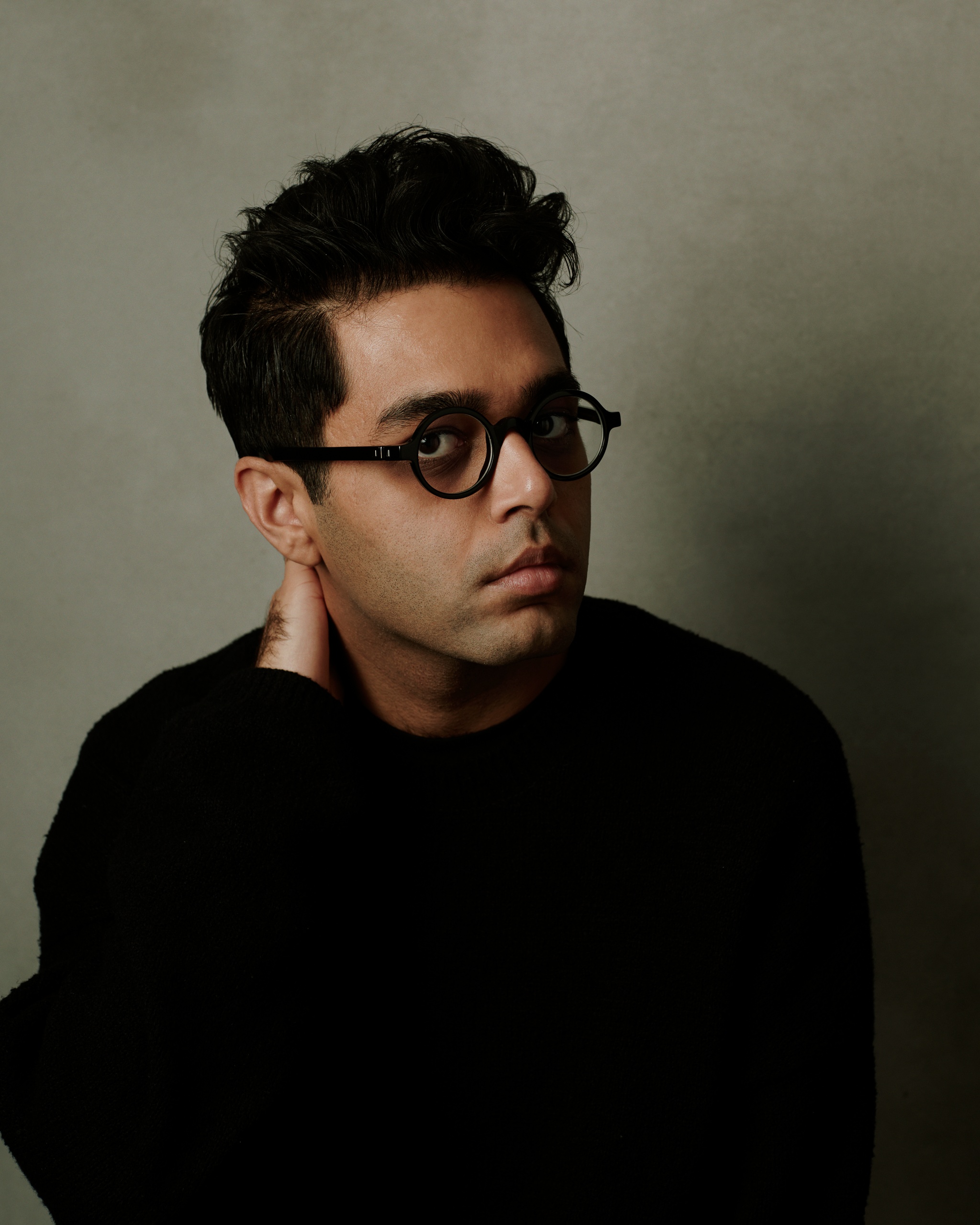 A portrait of Rafiq Bhatia who poses against a gray backdrop with one hand held behind his neck. Rafiq wears glasses and looks out of the side of them toward us. 