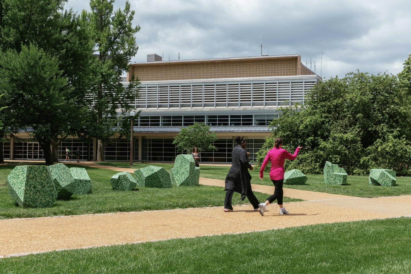 Two individuals walking on an outdoor path, past a series of 9 sculptural boulders in the grass. The scutpures are of various dimensions with planed faces; they are made of stainless steel-reinforced concrete with speckled light green glass mosaic tile.