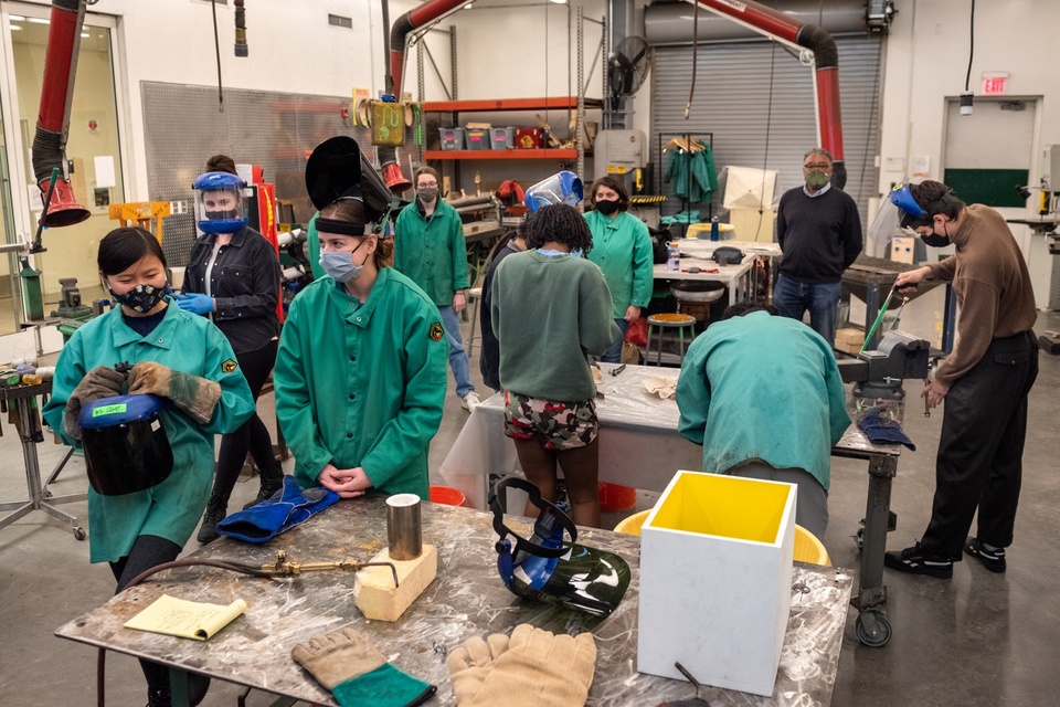 Wide angle of a metal shop full of people in protective gear doing various activities.