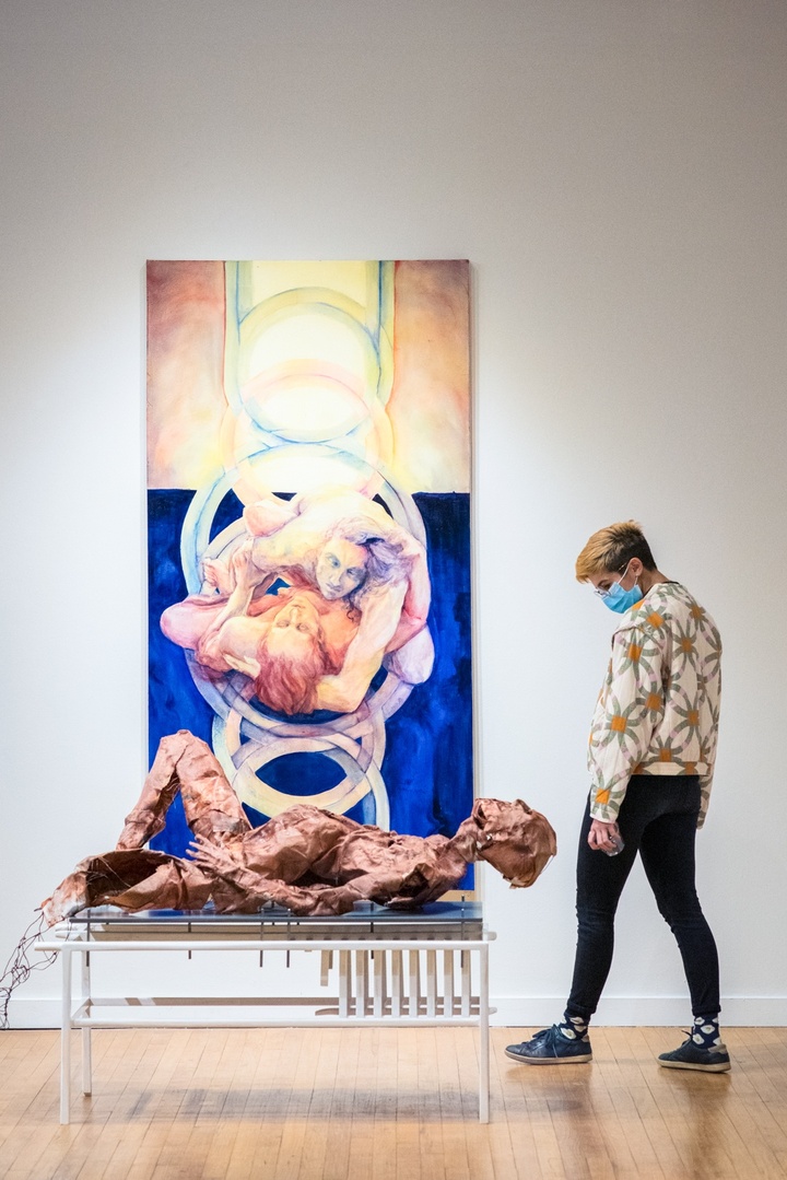 Person stands and looks at an installation which includes a vertical painting on the wall in deep blues and pinks of two bodies entwined. On a bench in front of it is a leathery mummified figure.