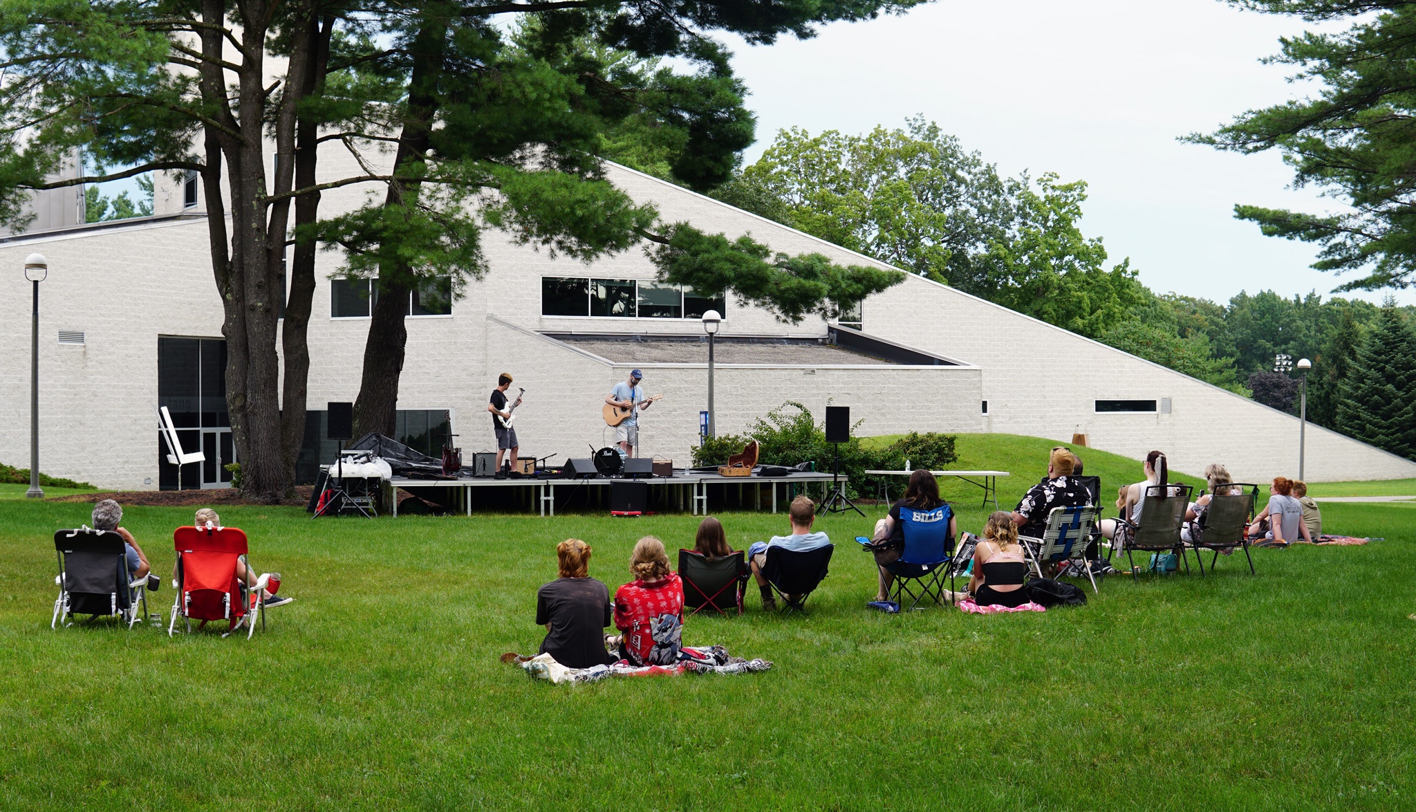 A small group of people sit on blankets and in lawn chairs in front of a small outdoor stage on which two people stand playing guitar.