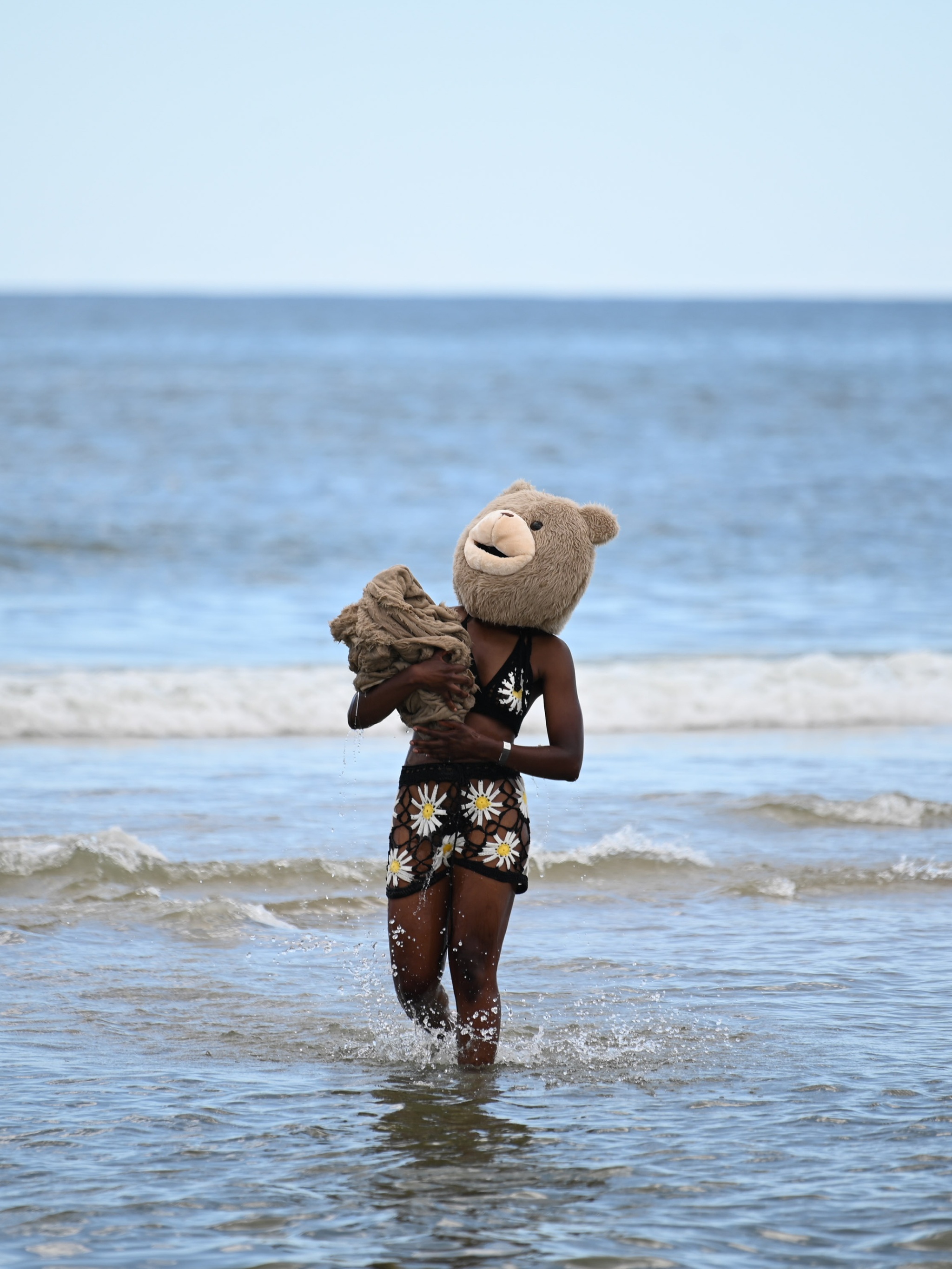 A Black person wearing a crocheted two-piece bathing suit with a yellow daisy design and a brown teddy bear costume head wades in the shallows of an ocean along a beach. They carry a bundle of brown fabric swaddled in their arms.