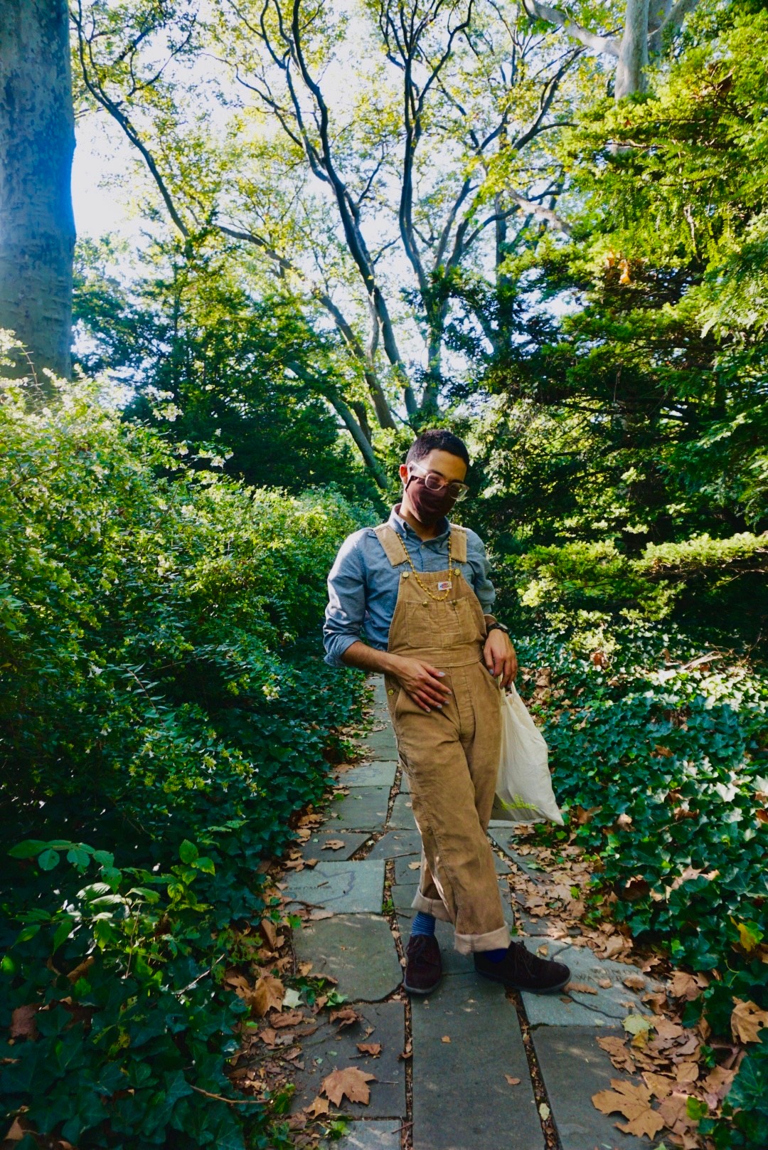 A queer brown person wearing tan courderoy overalls, mask, and pink glasses poses with one leg crossed over the other. They stand on a gray stone path littered with leaves and surrounded by greenery and branches. 