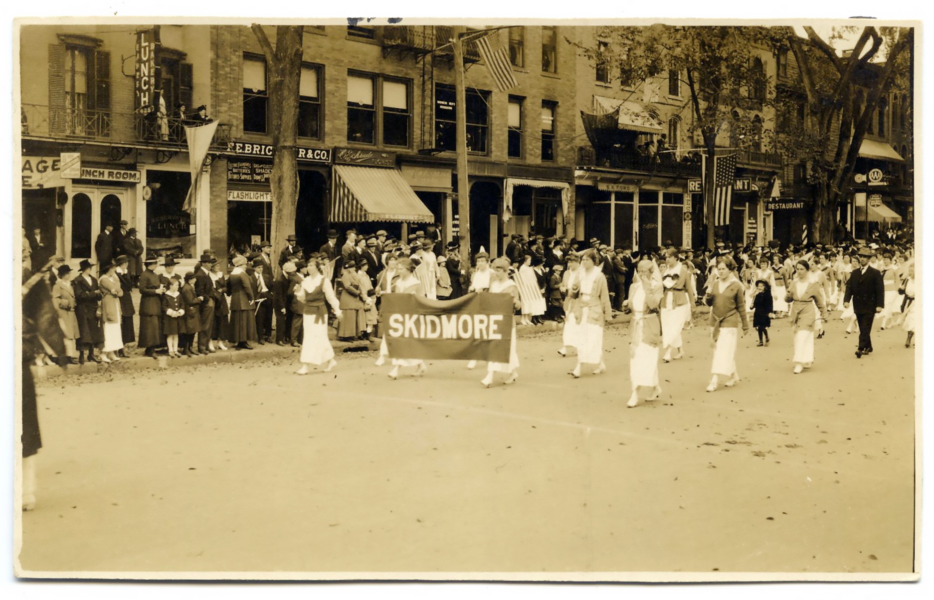 A sepia image depicts women in white dresses and darker coats parading on the street. Two women in the front are holding a banner saying “Skidmore.” Spectators witness the parade from the side.