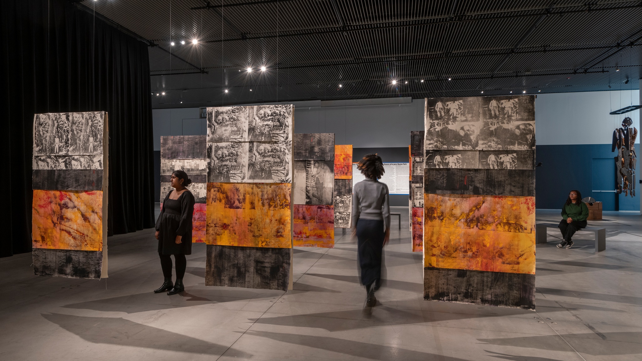 Nine painted fabric panels hang from a gallery ceiling in a grid. Two gallery visitors walk between the panels. The panels are painted in orange and black bands, with screen printed sections with black and white images of banana plantation workers in the 1920s.