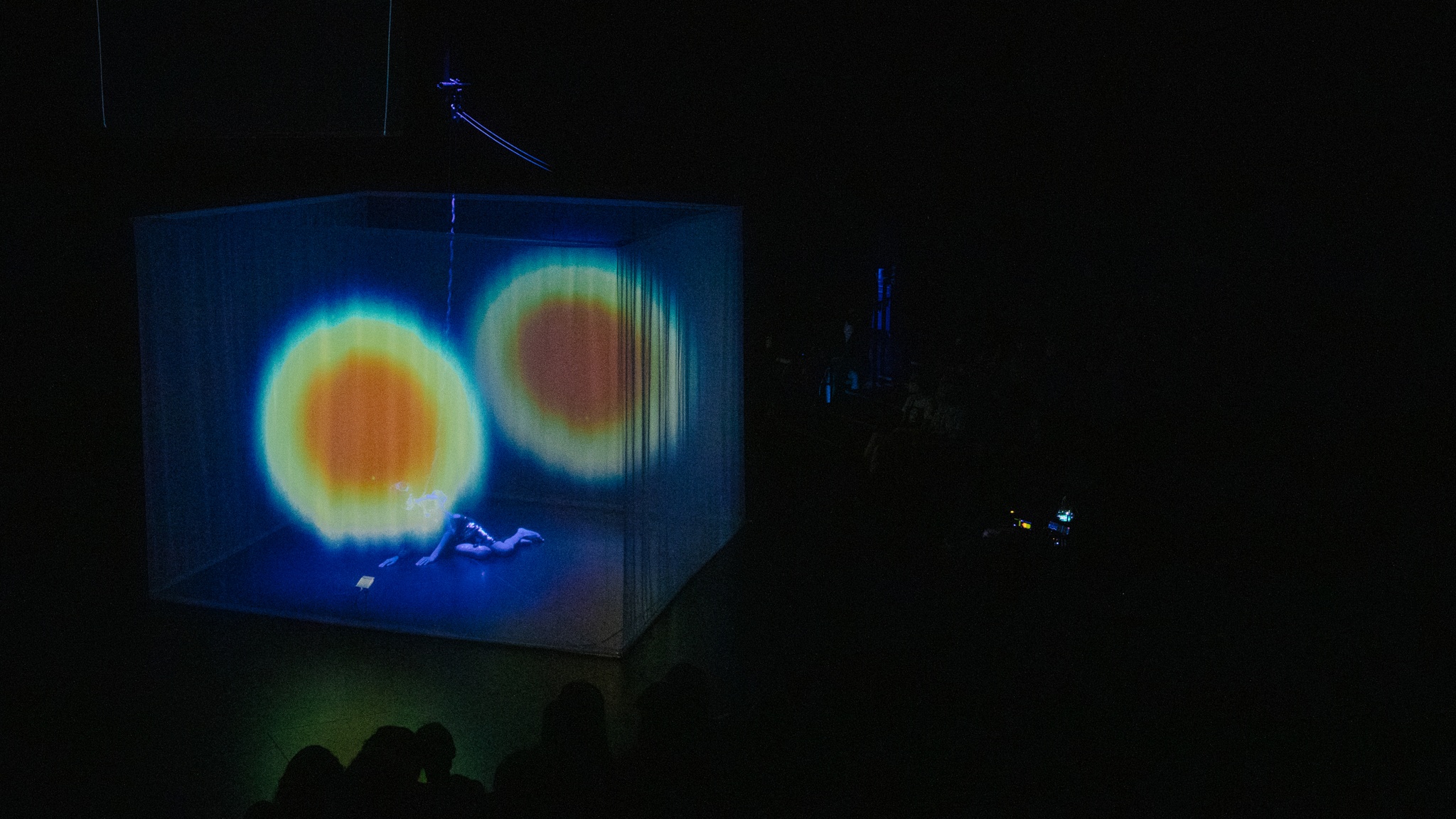 At the center of a darkened theater, sheer fabric hanging from the ceiling creates a cube on which a pulsating orange and green circle is projected. Through the fabric, a performer, the artist Yo-Yo Lin, can be seen wearing white lying on the floor.