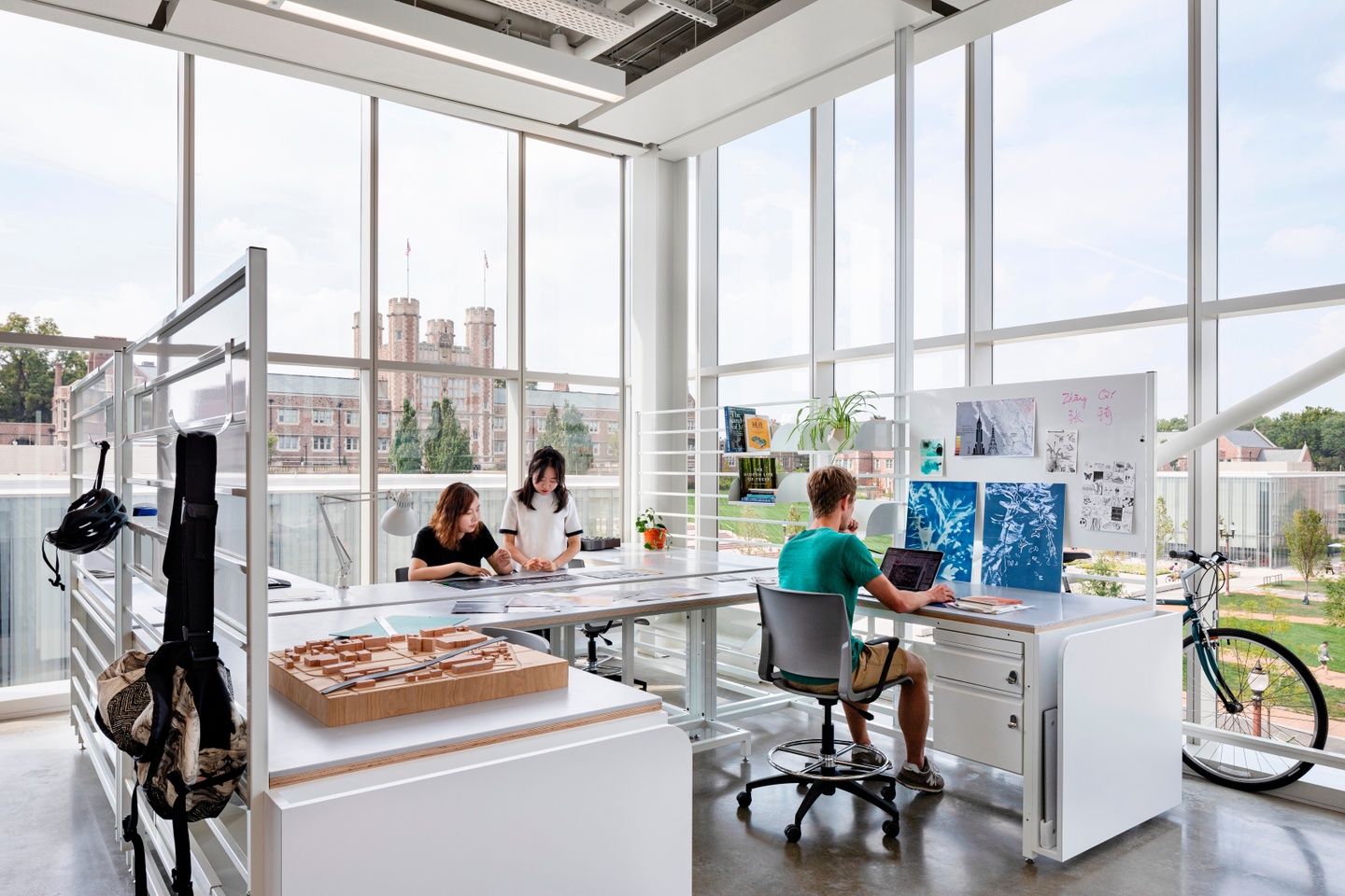 Three people work on their laptops at a modular, semi-private desk space to the backdrop of Brookings Hall, seen through the window wall.