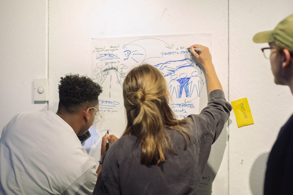 Two individuals write and draw on a paper posted up on the wall. The words Listen and Participate can be seen. Another individual looks toward them.