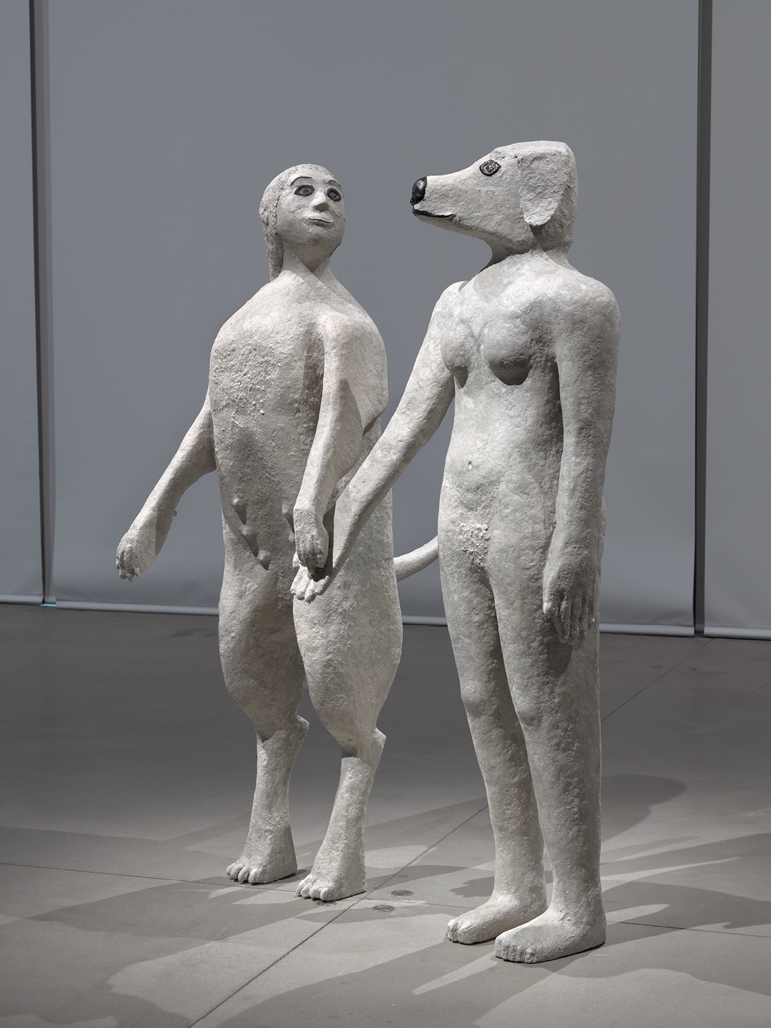 Two sculptures standing side by side. One is a woman with the body of a dog and the other is a dog with the body of a woman. They hold hands.