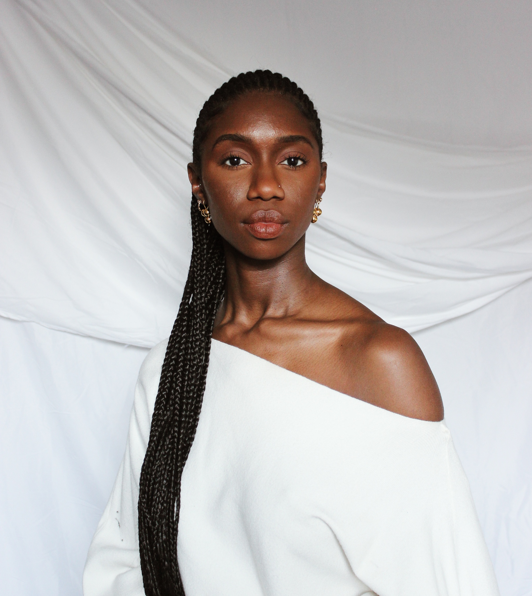 A portrait of Jenée-Daria Strand, a Guyanese American woman with dark brown skin. She wears a white dress with a bias cut neck that reveals one shoulder. Her long braids hang down over her other shoulder. She looks at us calmly without smiling. 