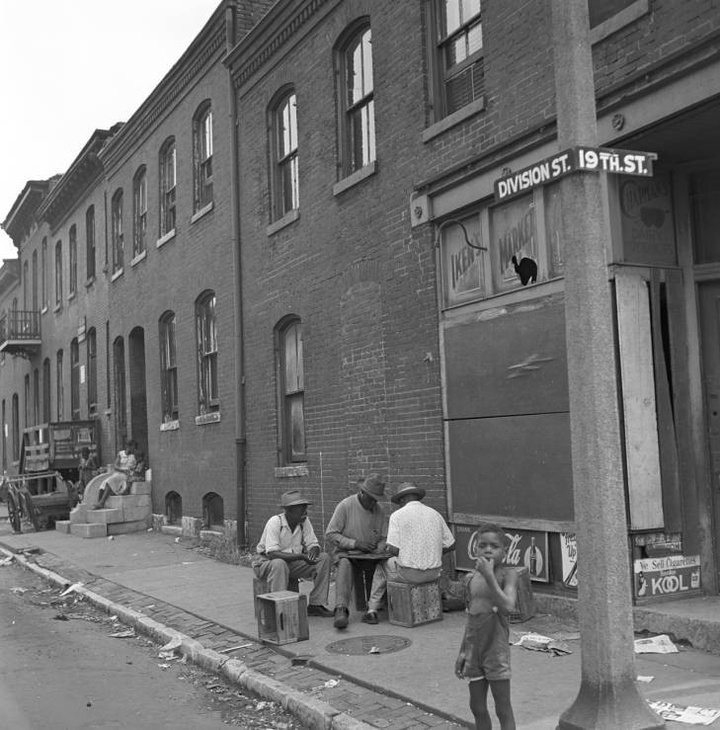 Black-and-white photo of a few people seated on crates outside brick buildings on a street corner, with a shirtlless child in the foreground near the light pole.