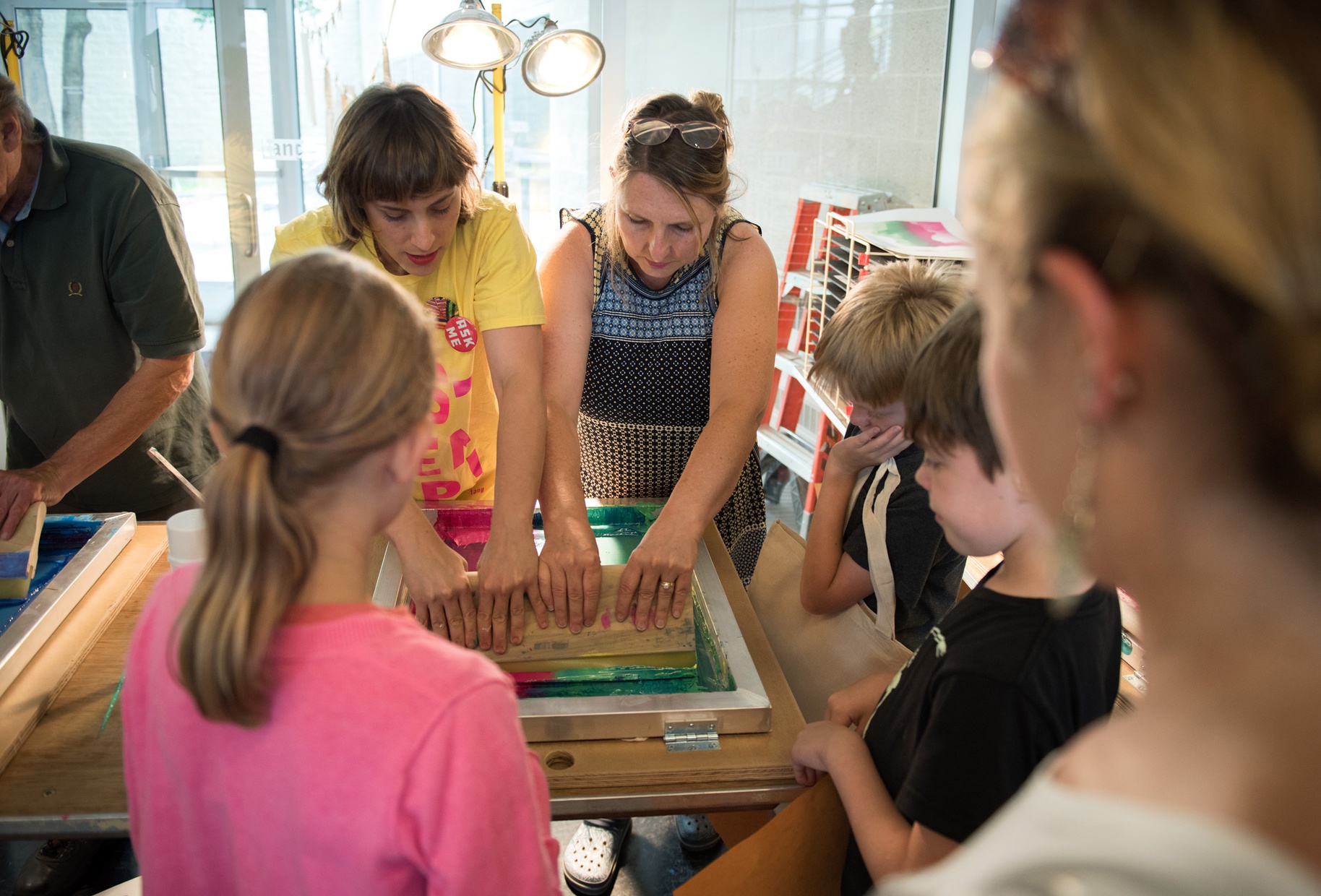 Two white women work on a screenprint while a group of children watch.