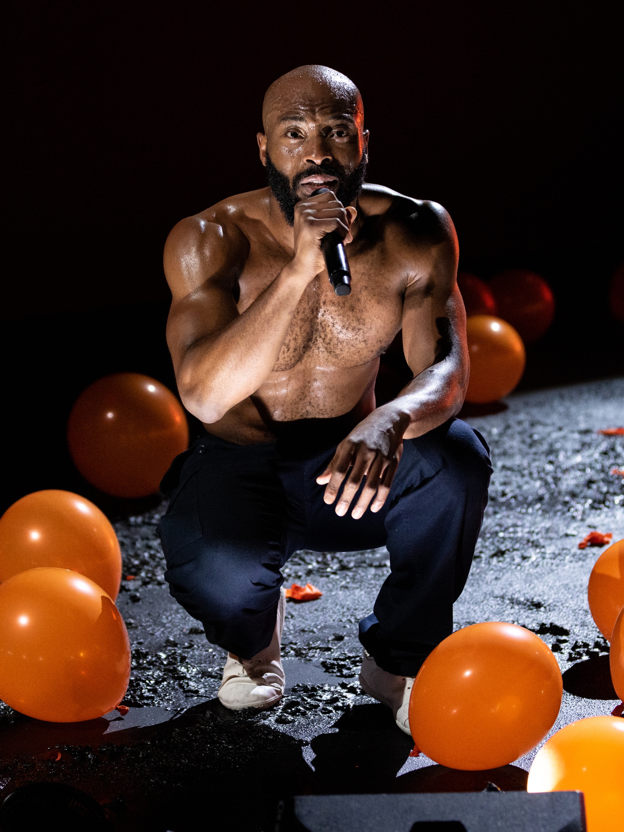 A Black man crouches on a dark stage lit dramatically with a bright light to the right. He is shirtless in blue pants with several inflated orange balloons drifting on the stage around him. He speaks into a microphone that he holds to his face. 