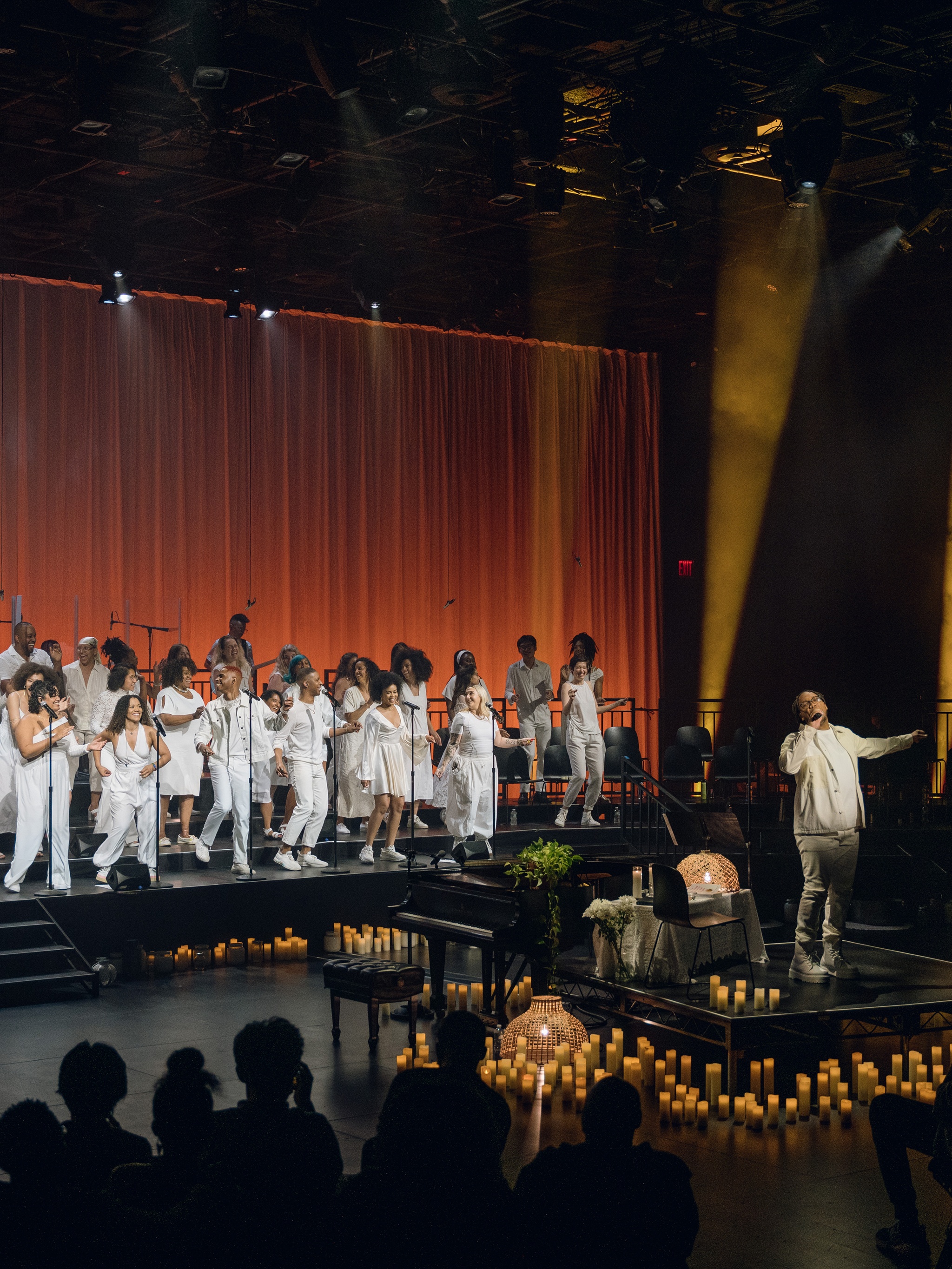 Troy Anthony, a Black man dressed in white, stands on a small stage in a black box theater with a choir on risers behind him. Troy stretches out his hands and the choir members dance. They are also dressed all in white. Around Troy are numerous lit candles. Behind the choir, a curtain is lit in deep umber tones.