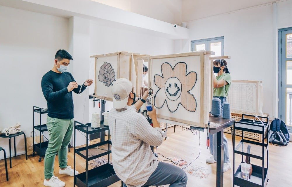 Two people in an open workshop space that are tufting on frames. One rug is a flower design.