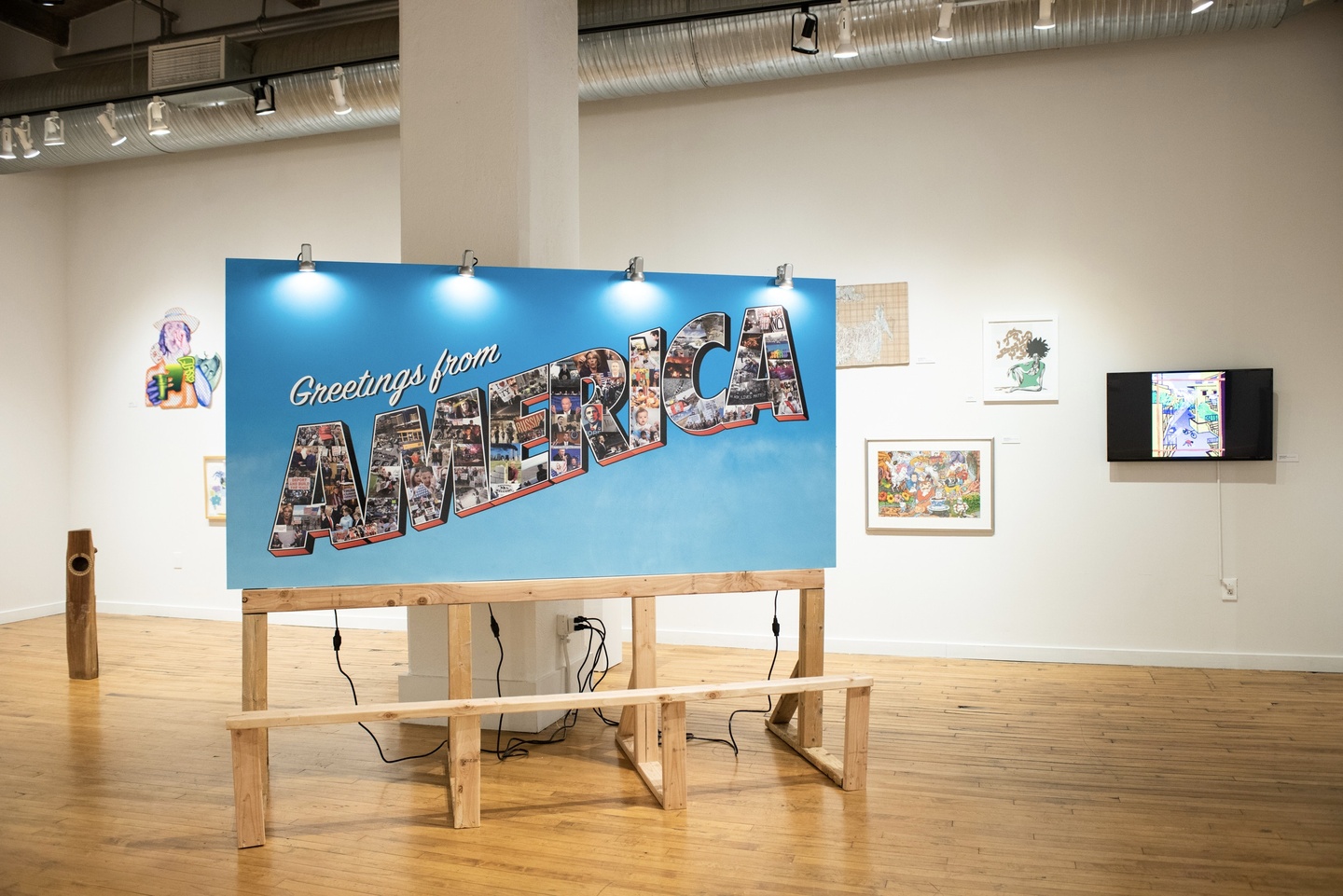 Large, horizontally oriened vinyl sign on MDF, with a bright blue background. White script type says "Greetings from" and the word AMERICA is in all caps with an outline, filled in with a variety of photos. In the background are other artworks hung on gallery walls.