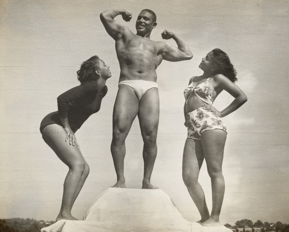 A black and white photograph of a muscular man flexing his arms while standing atop the middle of a three-tiered podium between two women in bathing suits smiling up at him.