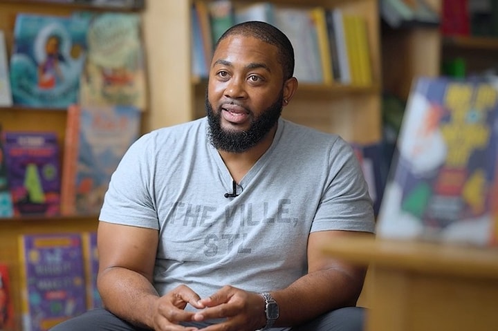 headshot of man with buzzed hair and a short beard in a grey shirt sitting in a studio with hands in lap
