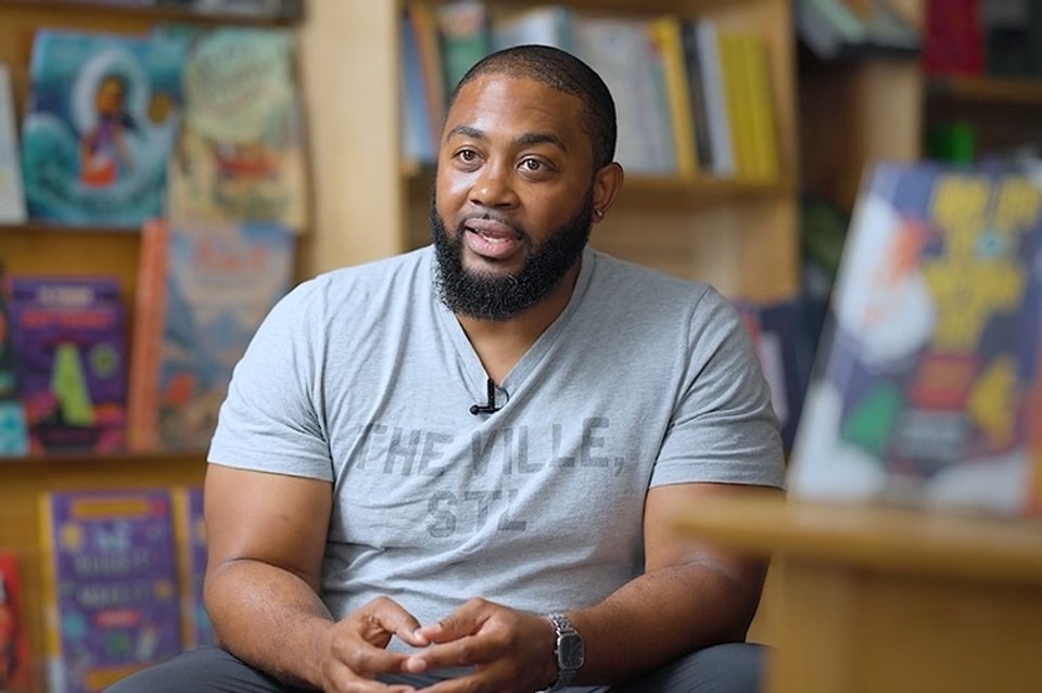 headshot of man with buzzed hair and a short beard in a grey shirt sitting in a studio with hands in lap