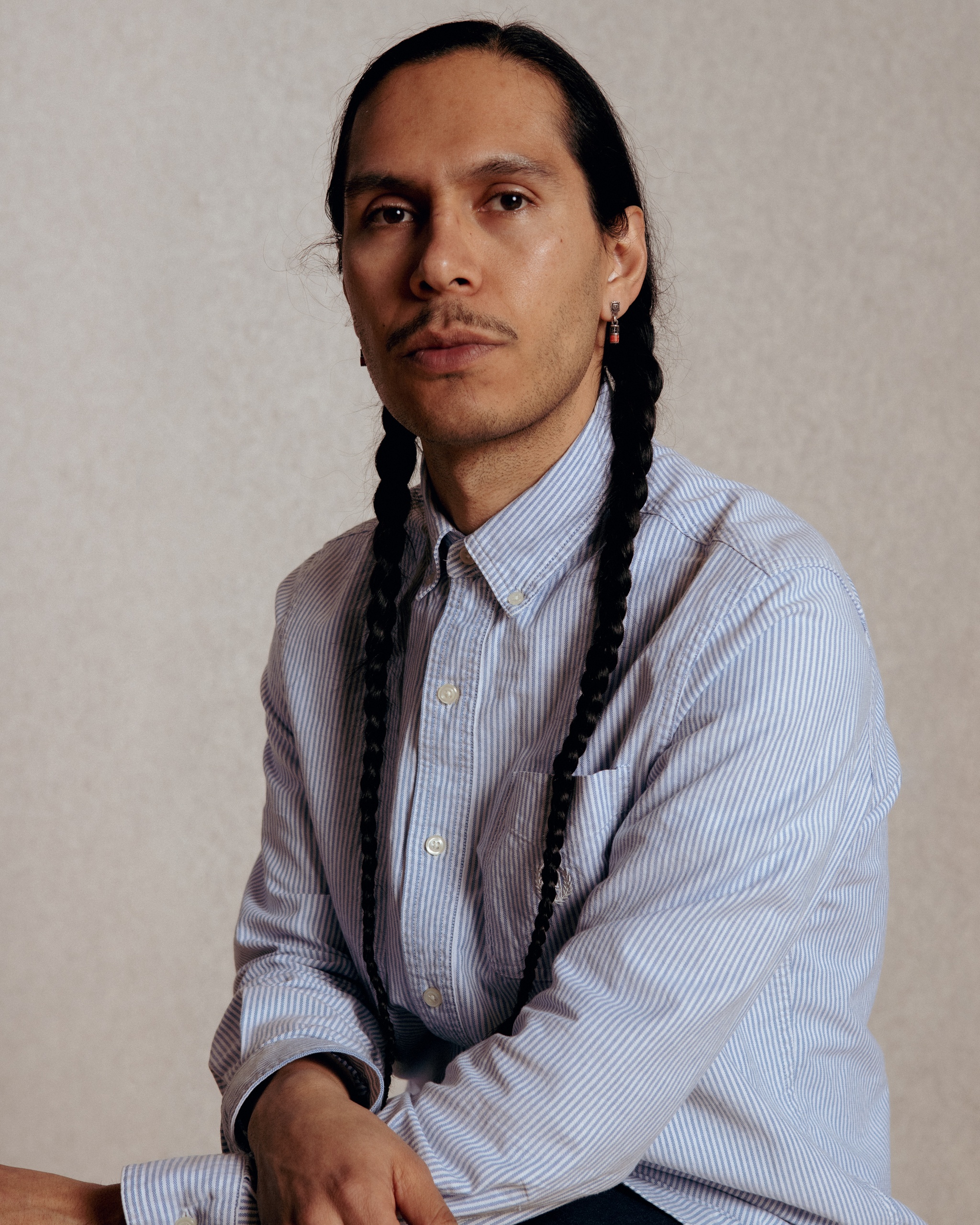 A portrait of artist Armando Guadalupe Cortés, a Mexican man who poses with arms crossed in his lap, facing us. Armando wears his dark hair in two long braids that hang down over either shoulder and wears a light blue button down shirt buttoned to the collar. Photo by Dana Golan 