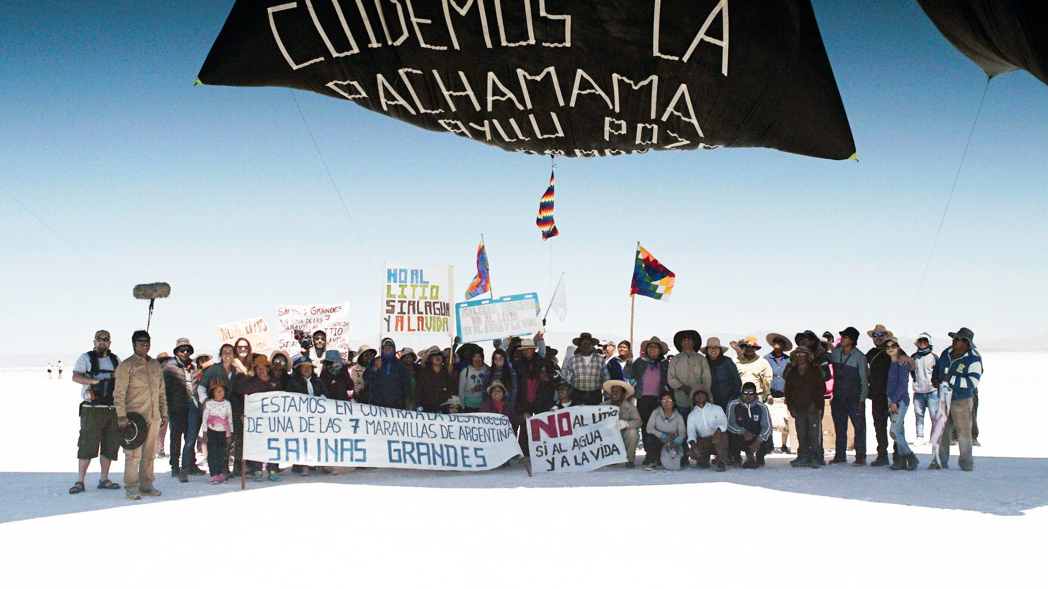A group of people gather with activist banners beneath a black hot air balloon on a white salt flat