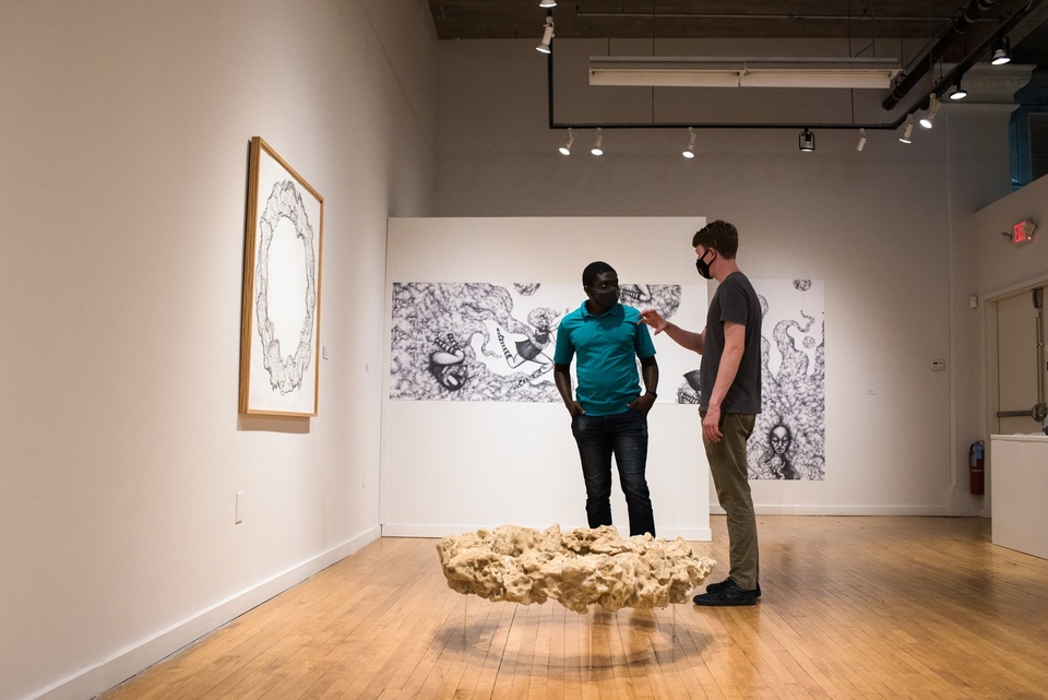 Two people in masks stand around a donut shaped object suspended a few inches off the floor of a gallery space and chat.