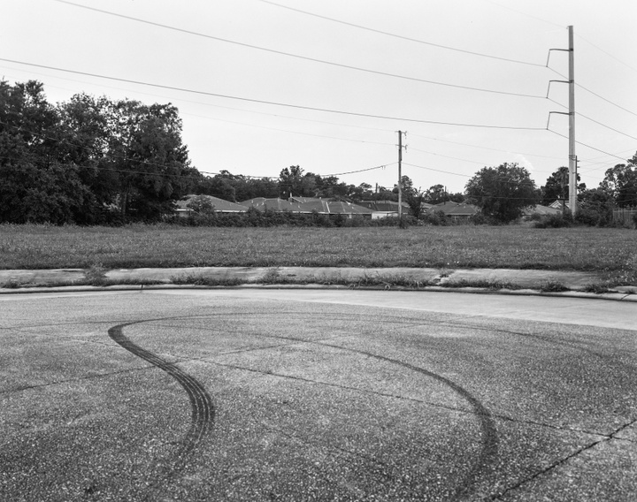 Black and white photograph of a road, houses and trees in the background. Skid marks in the foreground.