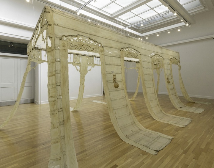 An intricate large off-white sculpture shaped like a building hanging from the ceiling in a gallery 