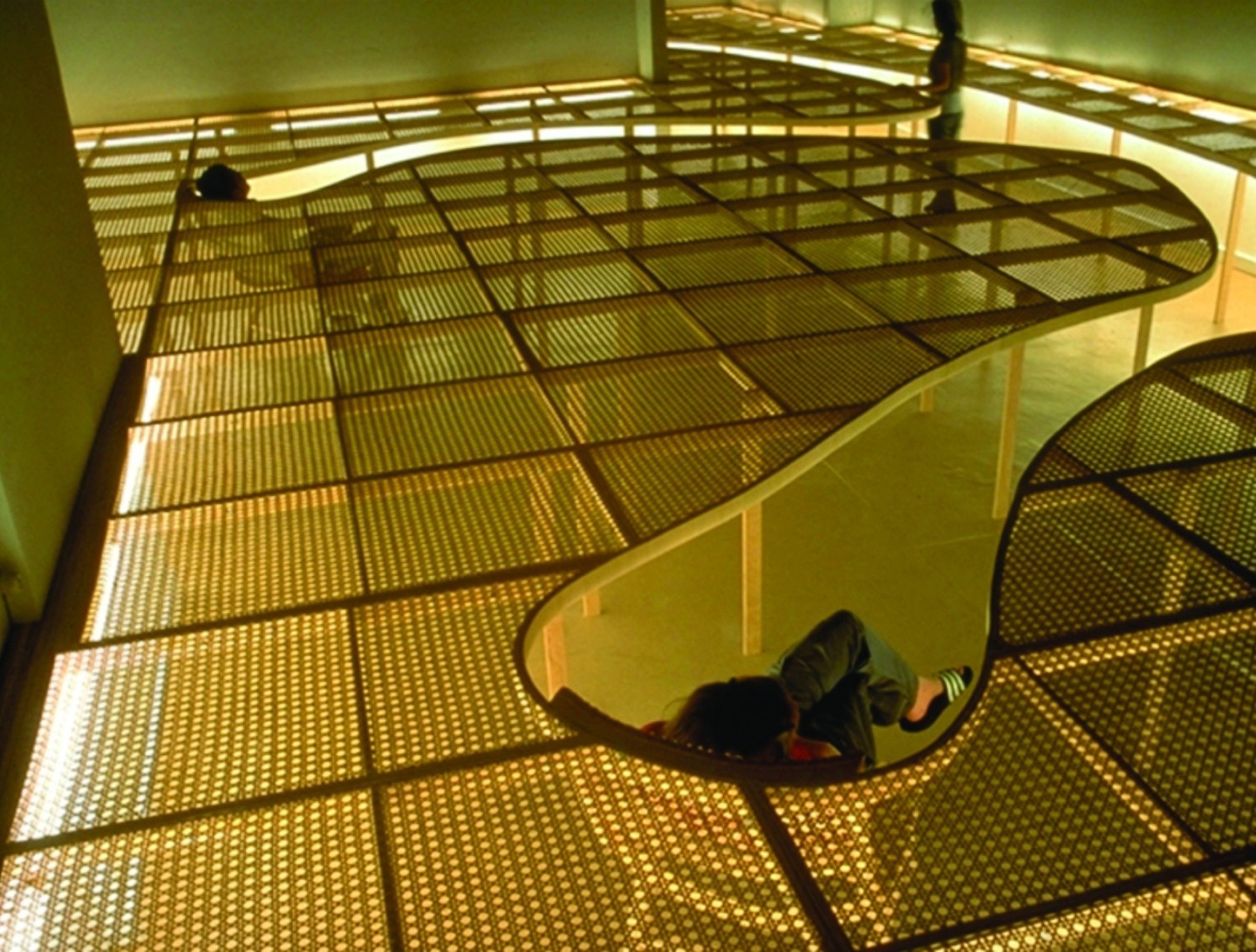 A dimly lit space, fluorescent lights illuminating the edge of a raised level of see-through architectural forms: two people sit in chairs below, as if sunken into the ground, so that they can see above the partitions into the vast space. Another person stands in the background.